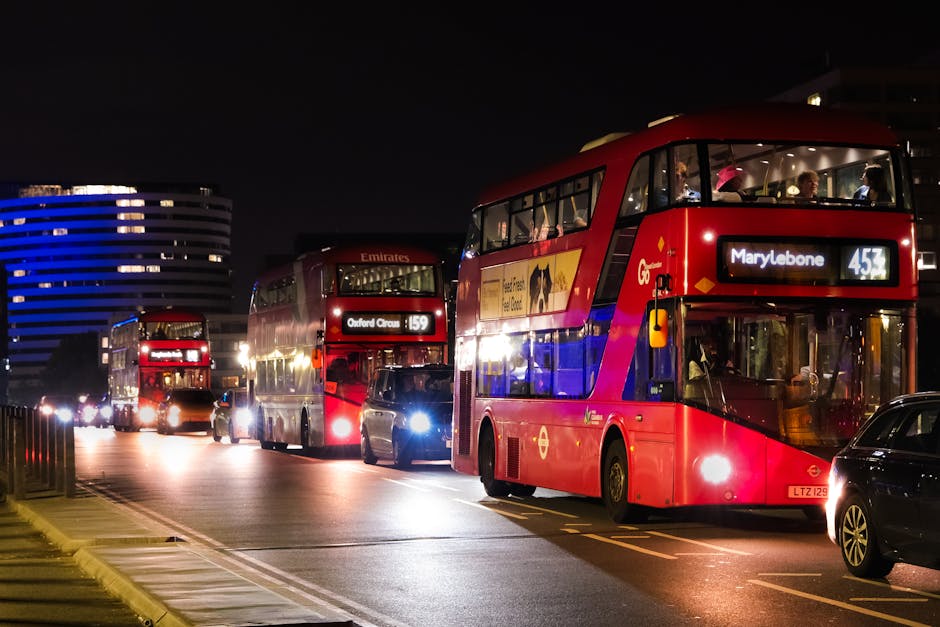 Nighttime street scene showing three red double-decker buses parked closely along the pavement, with illuminated interior lights visible through the windows. The bus in the foreground displays a route to Marylebone with the number 453, while another nearby bus shows a route to Oxford Circus with the number 159. Several cars are parked beside the buses, and a few vehicles with headlights on are visible along the street. In the background, a modern building with blue lighting is visible against a dark sky. The scene is well-lit by streetlights and vehicle headlights, and the area appears busy, indicative of urban transportation and home relocation activity. Man and Van Marylebone occasional mention is implied through the context of bus routes and urban setting related to removals and moving logistics.