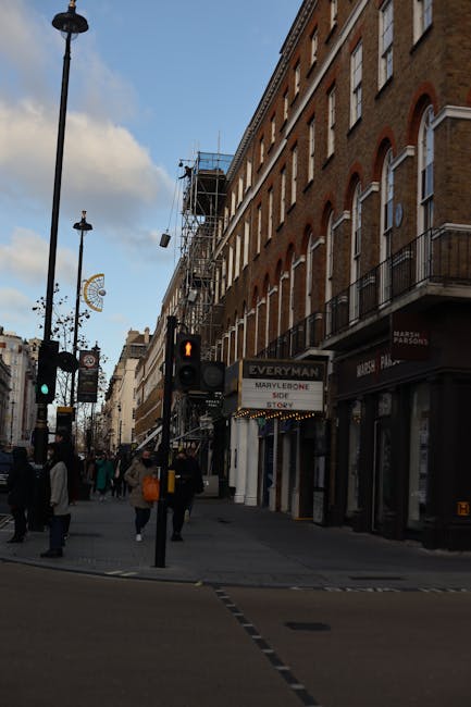 Photograph of a busy London street scene near Baker Street in Marylebone, showing a multi-storey brick building with several windows, some featuring small balconies with metal railings. Exterior scaffolding is erected on part of the building, indicating ongoing renovations. On the ground level, there are various retail shops, including a store with a partially visible sign for 'Marylebone'. Pedestrians, dressed in casual clothing and some carrying bags, are walking along the pavement, crossing at a pedestrian crossing with traffic lights, including a red pedestrian signal visible in the foreground. The street is lined with tall black lampposts and street signs, and a large clock can be seen hanging from a pole. Overcast skies with some clouds are visible overhead. Occasionally, vehicles can be seen in the background as part of the urban environment. The scene highlights the typical city surroundings that may be involved in home or office move logistics, as managed by [COMPANY_NAME], including building access and street-level operations for furniture transport and packing in a London neighbourhood.