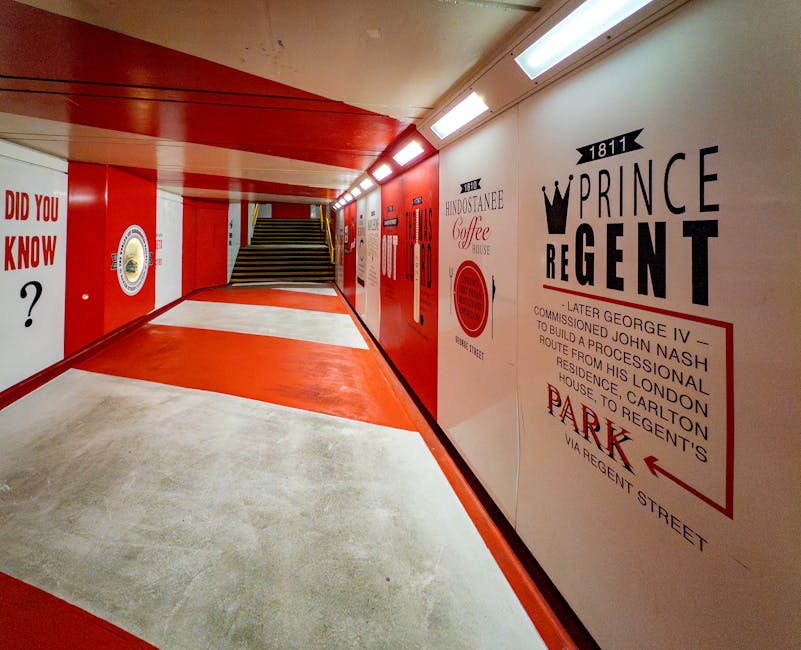 A photograph of a well-lit indoor pedestrian walkway with a red and white colour scheme, featuring a smooth concrete floor and a wooden panel ceiling. On the right wall, there are large graphic signs advertising Prince Regent and other local businesses, with text and a crown graphic. Moving further along, there is a staircase with black and yellow striped safety markings on the steps, leading upwards from the corridor. The left wall displays additional signage and artwork, including a partially visible question mark symbol and a circular emblem. The corridor appears clean and maintained, with no visible movement or objects being carried, and is likely part of a residential or commercial building, possibly during a house or office move as part of professional relocation services by Man and Van Marylebone.