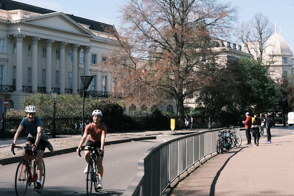 Photograph of two cyclists wearing helmets riding along a paved sidewalk adjacent to a road in Marylebone, with a black metal fence, bicycles parked against the fence, and people walking on the pavement. In the background, there is a large white building with classical architecture, columns, and decorative elements, alongside trees with sparse foliage indicating early spring or late autumn. The scene captures a typical urban street environment used for home relocation or moving logistics, with natural daylight and mild weather conditions. Man and Van Marylebone’s removal services may include navigating such city streets during furniture transport or home packing and moving activities, with equipment like loading trolleys or vehicle loading areas potentially involved in the process.