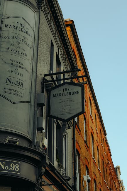 A close-up view of the exterior of a building housing The Marylebone pub, featuring a vintage-style black and white sign with text indicating the name, address on High Street, and offerings such as wines and spirits. The building has a curved corner with large windows, some with small balconies and plants, and a brick facade with visible window frames and decorative trim. The image is taken from a low angle against a clear blue sky, emphasizing the signage and architectural details, relevant to house removals and moving services provided by Man and Van Marylebone for logistics related to home relocation or furniture transport in the Marylebone area.
