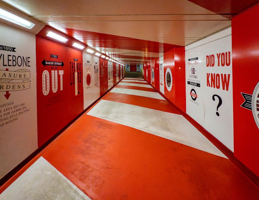 A long, narrow underground pedestrian corridor with a red and white colour scheme, featuring bold signage and directional arrows on the walls. The corridor has a low ceiling with evenly spaced rectangular lighting panels, illuminating the area evenly. The left wall displays various signs and posters, including the words 'LONGBOW' and 'OUT' with directional arrows, while the right wall features large red text reading 'DID YOU KNOW?' alongside other graphics and information. The floor is decorated with large, diagonal white and red sections, creating a striking visual pattern. The corridor appears to be part of a train station or underground passage, suitable for walking and light luggage movement, and could be used for planning or coordinating a home relocation or furniture transport as part of a professional moving service. Man and Van Marylebone may utilise such environments in their moving and packing logistics, with this setting representing an accessible, well-lit route for transporting boxes and furniture to a vehicle or different parts of a property.
