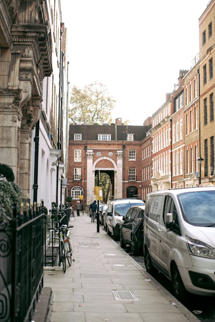A narrow city street in Marylebone with parked vehicles, including a white van and cars, lining the right side of the pavement. On the left, there are black metal railings and bicycles secured to them. The buildings on either side are multi-storey, made of brick and stone, with detailed architectural features such as decorative cornices and window frames. In the background, an arched brick gateway with white accents leads to a courtyard or another street, allowing visibility through to trees and additional buildings beyond. The pavement is clean and composed of rectangular stone slabs. The scene is illuminated by natural daylight, suitable for a home relocation or furniture transport process, which might involve an initial loading or unloading phase handled by Man and Van Marylebone for efficient house removals and packing services.