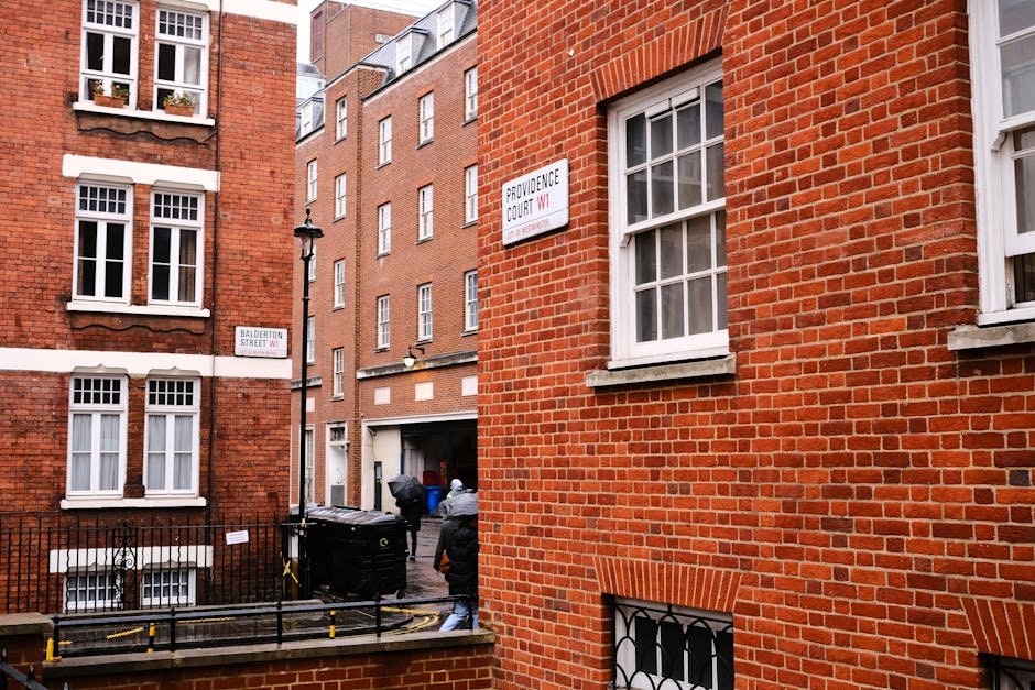 A view of a narrow urban street with red brick residential buildings on both sides, showing multiple white-framed sash windows and street signs indicating the intersection of Balderton Street W1 and Providence Court W1 in Marylebone. In the foreground, there is a black metal fence, a black wheelie bin, and a paved pavement area. Two men dressed in casual clothing and jackets are seen carrying or moving large cardboard boxes and wrapped furniture towards a small loading area at the rear of the buildings, which appears to provide access for a professional removals service such as Man and Van Marylebone. The scene is well-lit with natural daylight, capturing the process of home relocation, furniture transport, and packing for a house move amidst an urban environment consistent with successful loading operations for small apartment or townhouse moves.
