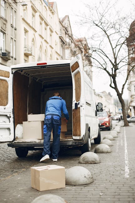A man in a blue jacket and dark trousers is loading large cardboard boxes and white furniture pieces into the open rear of a white commercial van parked on a city street. The van is positioned next to a curb with concrete bollards, and the surrounding environment features typical urban architecture with multi-storey buildings and leafless trees, suggesting late autumn or winter. The scene captures the loading process during daytime, with natural light illuminating the area, and indicates a home relocation or furniture transport operation. The interior of the van shows wooden paneling, and additional packing materials may be visible inside. This setting aligns with professional removals services like those offered by Man and Van Marylebone, supporting efficient packing and loading for house removals or same-day moving services.