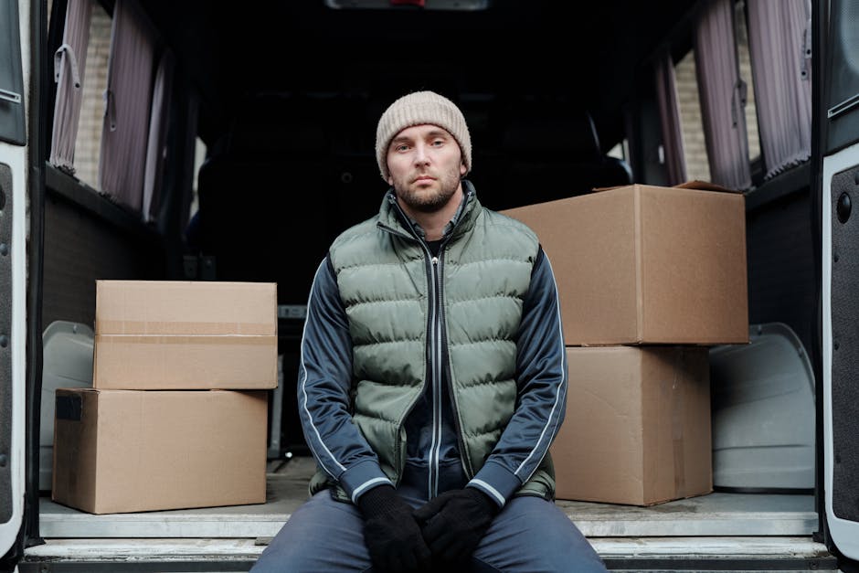 A young man wearing a beige knitted beanie, a green padded vest over a navy blue athletic jacket with white stripe details, and black gloves, sits on the open rear door sill of a moving van. Inside the van, several cardboard boxes of varying sizes are stacked on the floor, some with visible tape to secure the contents. The interior of the van shows black padded walls with vertical grooves, and the loading scene takes place on a paved or concrete surface, likely outside a residential property in Marylebone. The man appears to be involved in a home relocation or furniture transport process, with the boxes prepared for moving. The lighting is natural, and the scene suggests a carefully organized packing and loading process. The presence of the company Man and Van Marylebone is implied in the context of providing professional removals and urgent van services, supporting the subject of same-day removals in Marylebone.