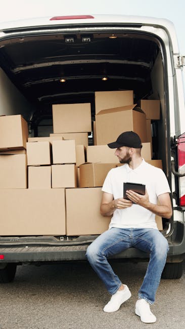 A man in a blue jacket and dark trousers is loading large cardboard boxes and white furniture pieces into the open rear of a white commercial van parked on a city street. The van is positioned next to a curb with concrete bollards, and the surrounding environment features typical urban architecture with multi-storey buildings and leafless trees, suggesting late autumn or winter. The scene captures the loading process during daytime, with natural light illuminating the area, and indicates a home relocation or furniture transport operation. The interior of the van shows wooden paneling, and additional packing materials may be visible inside. This setting aligns with professional removals services like those offered by Man and Van Marylebone, supporting efficient packing and loading for house removals or same-day moving services.