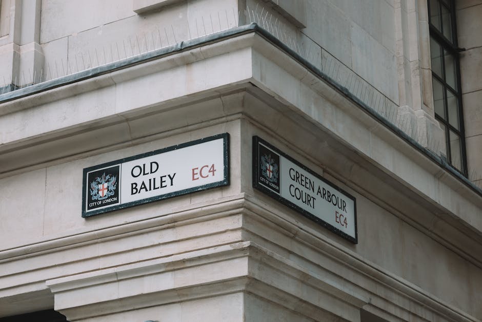 Photograph of the corner of a light-colored stone building at the intersection of Old Bailey and Green Arbour Court in the City of London, with two rectangular street signs affixed to the facade. The Old Bailey sign, on the left, displays the name 'OLD BAILEY' with the City of London crest and the postcode EC4 in red. The Green Arbour Court sign, on the right, shows the name 'GREEN ARBOUR COURT' along with the City of London crest and postcode EC4. The building features a decorative cornice and part of a window with black framing is visible above the signs. The scene is captured in natural daylight, emphasizing the urban environment. This setting relates to London street signage, which can be relevant in the context of house removals, packing, and moving logistics managed by companies like Man and Van Marylebone, especially around the historic City of London area.
