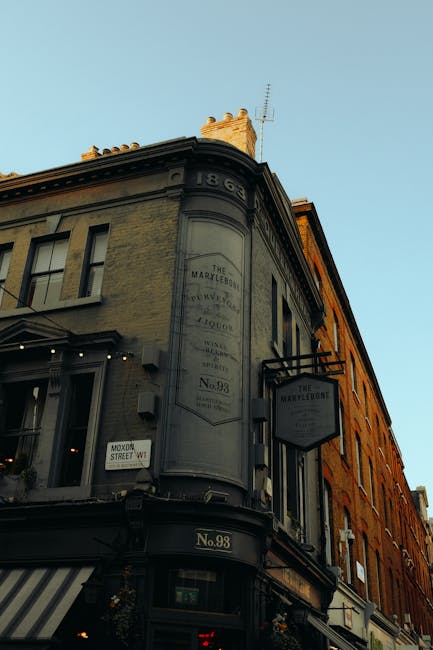A three-storey historic building with a rounded corner facade located on a street in Marylebone, London, captured during daylight with clear, blue sky. The building features a black and beige exterior with decorative signage for 'The Marylebone' pub or establishment, including vintage-style lettering and a hanging sign attached to the building's side. The ground floor has large windows with striped awnings, while the upper floors display multiple sash windows with white frames. The street scene suggests a busy area suitable for house removals or furniture transport, with visible signage and a street address number '98'. The image illustrates a typical setting for residential or commercial relocations in the Marylebone area, emphasizing the urban environment and traditional architecture of the neighborhood.