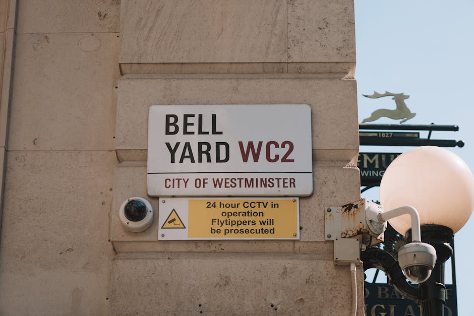 Close-up image of a building corner in Marylebone, Westminster, showing a white rectangular sign with black and red text indicating 'BELL YARD WC2' and 'CITY OF WESTMINSTER'. Below, a yellow sign with black text warns about 24-hour CCTV surveillance and that 'Flytippers will be prosecuted', attached to a beige stone facade. To the right, a spherical white street lamp with a black pole is visible, along with a security camera mounted on the wall near the signs. The background includes a clear blue sky and a partial view of a decorative metal sign with a leaping animal figure, suggesting urban street surroundings. This scene represents a typical location where house removals or furniture transport operations may occur, with infrastructure supporting logistics and security during home relocation services managed by Man and Van Marylebone.