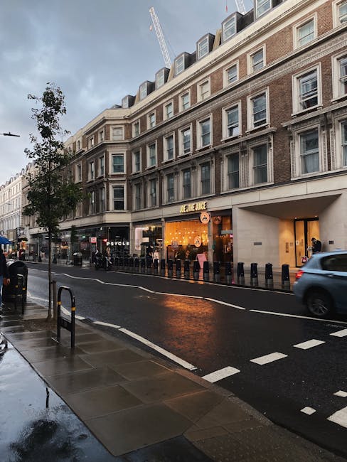 Photograph of a city street viewed from the sidewalk during a cloudy, rainy day, featuring wet pavement and puddles. A large multi-storey building with beige and white exterior walls and numerous windows lines the street. On the ground floor, a furniture and home accessories store with a brightly lit interior window display is visible, displaying lamps, decorative items, and seating furniture. To the left of the store, a black bicycle is parked against a tree, and several pedestrians are walking along the sidewalk. On the road, a grey car is passing by, and some bollards separate the parking area from the main road. A construction crane is visible in the background above the building, indicating ongoing building work. The scene reflects urban activity and is relevant to logistical aspects of home relocation and furniture transport, with the presence of a moving service provider like Man and Van Marylebone potentially involved in the logistics of house removals on Wigmore Street.